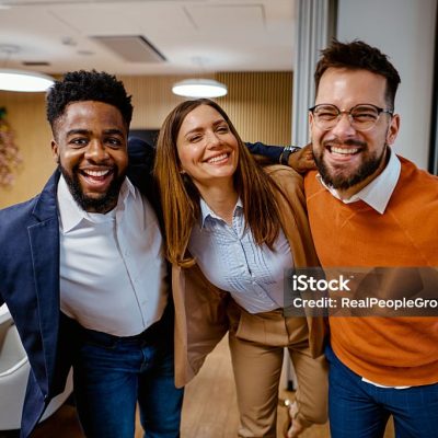 Three cheerful business colleagues are standing together with arms around each other, smiling and looking at the camera, showing thumbs up, enjoying their teamwork in a modern office