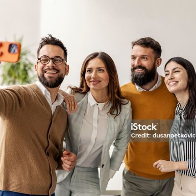 Close-up portrait of mid-aged diverse professionals in business casual attire taking group selfie in bright modern office. Natural lighting highlights casual corporate environment and team camaraderie.